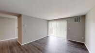 This single-floor unit offers a thoughtfully designed layout encompassing a combined kitchen and living room, one bedroom, one bathroom, a dedicated closet, and a hallway connecting the spaces. The living room is a bright, open area finished with dark hardwood-style flooring and neutral gray walls. A large sliding glass door fitted with vertical blinds serves as the room's focal point, allowing generous natural light and providing outdoor access. A ceiling fan is visible from the hallway entry, adding both function and subtle character.

The kitchen is efficiently arranged in an L-shaped configuration, featuring dark cabinetry, light countertops, a subway tile backsplash, and stainless steel appliances including a gas range, dishwasher, and over-range microwave. A window above the sink brings in additional natural light. The kitchen transitions seamlessly into the living area via light-toned hardwood-style flooring.

The bedroom is a well-sized, carpeted room finished in the same neutral gray palette. It includes access to a dedicated walk-in closet with built-in shelving and a direct connection to the hallway. The bathroom features a full tub-shower combination with striking vertically-laid gray tile surround, wood-look tile flooring, a white vanity, and brushed nickel fixtures, delivering a clean, contemporary aesthetic throughout.