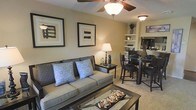 This two-floor residence offers a thoughtfully designed interior that balances comfort and style. The main living area features a warm, neutral color palette with beige walls that create an inviting atmosphere. A ceiling fan with a central light fixture provides both airflow and ambient lighting, complementing the soft carpeted flooring that runs throughout the space.

The living room is anchored by a cushioned sofa accented with patterned and blue decorative pillows, paired with a glass-top coffee table. Framed abstract artwork adorns the walls, adding a contemporary touch. A side table with a classic table lamp provides additional warm lighting to the seating area.

The dining area flows seamlessly from the living room, featuring a dark-toned dining table and matching chairs, creating a cohesive and elegant contrast against the lighter surroundings. Built-in shelving along the wall offers display space for decorative accents, adding character to the open-plan layout.

The open-concept design between the living and dining areas enhances the sense of space and promotes an easy, functional flow. The overall ambiance is warm, composed, and residential in character, making it a comfortable and visually appealing multi-level home.