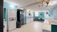 This shared laundry facility is a functional and practical space designed to accommodate a high volume of laundry needs. The room is immediately recognizable by its distinctive teal-painted walls, which give the space a consistent and identifiable character throughout. The layout is thoughtfully organized, with a generous row of front-loading washing machines lining one side and a combination of stacked and individual dryers positioned along the opposite wall, providing ample capacity for simultaneous use. A centrally located vending machine offers added convenience for users during their visit. The light-tiled flooring is easy to maintain and contributes to the room's clean, utilitarian appearance. Exposed overhead piping runs across the textured ceiling, reflecting the practical nature of the space, while track lighting ensures the room is well-lit throughout. A small seating area with a round table and chairs is tucked near the windows, allowing users to wait comfortably while their laundry cycles complete. The windows, fitted with security grilles, admit natural light and contribute to ventilation alongside a wall-mounted fan. A wall-mounted monitor and clearly posted informational signage further enhance the usability and organization of the facility. Fire safety equipment, including a sprinkler head, is also present, underscoring attention to safety standards.