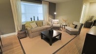 This single-floor space offers a refined and welcoming atmosphere, anchored by a tastefully designed living area. The room showcases a neutral color palette of warm beiges, soft grays, and earthy tones, complemented by a bold dark accent wall that adds depth and contrast to the overall aesthetic. A large bay window fitted with white blinds and flowing sheer curtains floods the space with abundant natural light, creating an open and airy feel. The living area is centered around a substantial dark espresso square coffee table, which grounds the room and provides a striking visual focal point against the light-toned area rug with a subtle border detail. Seating is generously arranged and includes a plush slipcovered sofa adorned with green and neutral accent pillows, a classic armchair, and a sleek chaise lounge, offering ample comfort for relaxing or entertaining. Decorative touches such as a tall orchid arrangement in a dark ceramic pot, a floral vase, and a minimalist arc floor lamp add elegance and personality to the space. The hardwood flooring visible at the room's edges provides warmth and a natural finish. Overall, the space presents a calm, sophisticated ambiance ideal for comfortable everyday living.