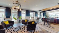 This single-floor clubhouse is thoughtfully designed to offer a variety of social and recreational spaces, all unified by a bold and contemporary aesthetic. Deep navy walls paired with white shiplap wainscoting create a striking visual contrast throughout, complemented by stone-tile flooring in the main lounge and dining areas.

The main lounge area serves as a welcoming gathering space, featuring plush charcoal armchairs with yellow accent pillows arranged atop a bold black-and-white spotted area rug. An eye-catching orb chandelier with exposed Edison-style bulbs anchors the room, while large multi-pane windows flood the space with natural light and offer views of the surrounding greenery. A built-in work counter along the windowed wall provides a functional workspace.

Adjacent to the lounge, the bar area features a dark countertop island with bold red barstools, patterned accent wallpaper, pendant lighting, and an under-counter beverage refrigerator — ideal for entertaining. High-top tables with sleek bar-height chairs offer additional seating nearby.

A separate game room provides a distinct change in atmosphere, with patterned carpeting, moody gray walls, and a central pool table beneath a modern brass chandelier. A vibrant teal sectional sofa, yellow accent ottomans, wall-mounted artwork, and a large flat-screen TV complete this energetic entertainment space.