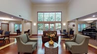 This single-floor community space is thoughtfully designed to blend elegance with functionality across several distinct areas. The grand entrance lobby immediately impresses with soaring double-height ceilings, a sweeping wall of large windows and glass doors that flood the space with natural light and frame lush outdoor greenery. A tasteful seating arrangement with upholstered chairs and a decorative coffee table anchors this welcoming area.

Adjacent to the lobby, a spacious common lounge features rich hardwood flooring transitioning to plush carpeting, decorative columns, and a classic fireplace with a detailed mantel and mirror above. Built-in bookshelves flank the hearth, while a baby grand piano adds a refined, cultural focal point. A wall-mounted television and additional seating areas make the lounge versatile for both relaxation and social gatherings.

The dining area accommodates multiple tables and chairs finished in warm wood tones, with pendant lighting and windows offering outdoor views. A dedicated beverage station includes generous cabinetry with granite-style countertops and a coffee maker, providing a convenient self-service refreshment area. Throughout, consistent hardwood flooring, decorative columns, and warm neutral tones create a cohesive, inviting ambiance.