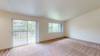 This single-floor residence offers a well-organized layout with a clean, neutral aesthetic throughout. The spacious living area is a standout feature, highlighted by vaulted ceilings, plush wall-to-wall carpeting, and an abundance of natural light streaming in through large windows and a sliding glass door that opens to a private balcony overlooking mature, verdant landscaping. The open sightlines between the living area and dining space create a comfortable, connected flow, with the dining area accented by a classic chandelier. The galley-style kitchen is efficiently designed with white cabinetry, ample counter space, light wood-look flooring, a double sink, and white appliances including a refrigerator and electric range. A dedicated in-unit laundry area provides added convenience with full-size washer and dryer connections and overhead shelving. The bedrooms feature plush carpeting and sliding-door closets with built-in hanging rods. Two bathrooms serve the unit: a full bathroom with a single vanity, large mirror, and soaking tub, and a second bathroom equipped with a walk-in shower featuring sliding glass doors. Light wood-look flooring carries through the kitchen, bathrooms, and laundry area, providing a cohesive and easy-to-maintain finish throughout the non-carpeted spaces.