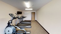 This single-level fitness room offers a functional and well-organized space for exercise. The room features neutral-toned walls and patterned carpet flooring, creating a clean and practical environment. Overhead fluorescent lighting ensures consistent illumination throughout the space, while large sliding glass doors along one wall invite abundant natural light and provide views of the surrounding outdoor area.

The room is equipped with a variety of exercise machines, including multiple treadmills, an elliptical trainer, a multi-function cable weight machine, and a rowing-style machine, providing options for both cardiovascular and strength training workouts. A wall-mounted flat-screen television is positioned for easy viewing during exercise sessions.

Climate comfort is addressed with a wall-mounted mini-split HVAC unit, allowing for independent temperature control within the space. A standard interior door provides access to the broader building. The overall layout is straightforward and efficiently arranged, maximizing usable floor space while keeping equipment accessible. The combination of natural light, practical amenities, and a varied selection of equipment makes this a well-rounded fitness facility suited for a range of workout preferences.