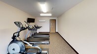 This single-level fitness room offers a functional and well-organized space for exercise. The room features neutral-toned walls and patterned carpet flooring, creating a clean and practical environment. Overhead fluorescent lighting ensures consistent illumination throughout the space, while large sliding glass doors along one wall invite abundant natural light and provide views of the surrounding outdoor area.

The room is equipped with a variety of exercise machines, including multiple treadmills, an elliptical trainer, a multi-function cable weight machine, and a rowing-style machine, providing options for both cardiovascular and strength training workouts. A wall-mounted flat-screen television is positioned for easy viewing during exercise sessions.

Climate comfort is addressed with a wall-mounted mini-split HVAC unit, allowing for independent temperature control within the space. A standard interior door provides access to the broader building. The overall layout is straightforward and efficiently arranged, maximizing usable floor space while keeping equipment accessible. The combination of natural light, practical amenities, and a varied selection of equipment makes this a well-rounded fitness facility suited for a range of workout preferences.