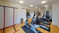 Located at 26 Haven St in Reading, MA, this dedicated fitness center offers a clean, well-organized workout environment spread across a single floor. The space features warm hardwood flooring throughout, complemented by crisp white walls and flush-mounted ceiling lights that create a bright and inviting atmosphere. A large window at one end of the room allows natural light to filter in, providing views of the surrounding neighborhood and adding an airy quality to the space.

The cardio section is well-equipped with multiple elliptical trainers and a stationary bike, each placed on protective rubber mats to preserve the hardwood floor beneath. A freestanding pull-up and dip station in red adds a functional strength-training option without requiring additional floor anchoring. On the opposite side of the room, a multi-function cable and weight machine offers a comprehensive strength-training setup, accompanied by a flat bench, catering to a range of resistance exercises.

A wall-mounted split air conditioning unit ensures comfortable year-round climate control. Large mirrors along one wall help visually expand the space while allowing users to monitor their form during workouts. A glass-paneled interior door provides easy access to and from the room. The overall design is practical and efficient, prioritizing functionality while maintaining a tidy, professional aesthetic suited for regular fitness use.