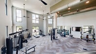 Located at 700 SE Veranda Pl in Port Saint Lucie, Florida, this two-floor fitness facility offers a thoughtfully designed environment for a comprehensive workout experience. The ground floor houses a generously proportioned exercise room defined by soaring double-height ceilings and large windows that flood the space with natural light and outdoor views. This level features a dedicated strength and cardio zone outfitted with treadmills, ellipticals, rowing machines, cable machines, benches, and an array of free weights and medicine balls. Herringbone-patterned carpet tiles cover the exercise floor, lending a modern, refined aesthetic, while exposed industrial ductwork running overhead adds an urban character to the space. A prominent staircase with sleek metal railings connects to the upper level. Floor-to-ceiling mirrors visually expand the space and provide functional feedback during workouts. The upper floor transitions into an open, airy studio space with light hardwood flooring, shiplap-clad columns, additional mirrored walls, and seamless glass railings overlooking the floor below. This level also includes a living area and provides direct outdoor access. Throughout both floors, recessed lighting and ceiling fans ensure a comfortable and well-lit environment suited for a full range of fitness activities.