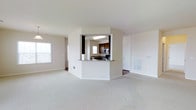 This single-floor residential space offers a clean, functional layout designed for comfortable everyday living. The open-concept living area is expansive and filled with natural light from multiple windows, featuring neutral-toned wall-to-wall carpeting that adds warmth and cohesion throughout the space.

A defining architectural feature is the partial partition wall that frames the kitchen, creating a semi-enclosed pass-through design. This setup visually connects the kitchen to the living area while maintaining a sense of defined space. The kitchen is equipped with dark espresso-toned cabinetry, stone-look granite countertops, and stainless steel appliances including a microwave and range. A built-in ceiling light fixture illuminates the kitchen area, and a window above the counter brings in additional natural light.

The transition between the carpeted living space and the hard-flooring kitchen is clearly delineated, adding visual contrast to the interior. A paneled interior door is visible from the main living area, suggesting access to additional rooms or storage. The hallway leading further into the unit hints at additional private spaces beyond the main living zone.

Overall, the space presents a straightforward, well-lit residential layout with practical design choices, neutral finishes, and a cohesive color palette of whites, beiges, and dark wood tones.