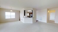 This single-floor residential space offers a clean, functional layout designed for comfortable everyday living. The open-concept living area is expansive and filled with natural light from multiple windows, featuring neutral-toned wall-to-wall carpeting that adds warmth and cohesion throughout the space.

A defining architectural feature is the partial partition wall that frames the kitchen, creating a semi-enclosed pass-through design. This setup visually connects the kitchen to the living area while maintaining a sense of defined space. The kitchen is equipped with dark espresso-toned cabinetry, stone-look granite countertops, and stainless steel appliances including a microwave and range. A built-in ceiling light fixture illuminates the kitchen area, and a window above the counter brings in additional natural light.

The transition between the carpeted living space and the hard-flooring kitchen is clearly delineated, adding visual contrast to the interior. A paneled interior door is visible from the main living area, suggesting access to additional rooms or storage. The hallway leading further into the unit hints at additional private spaces beyond the main living zone.

Overall, the space presents a straightforward, well-lit residential layout with practical design choices, neutral finishes, and a cohesive color palette of whites, beiges, and dark wood tones.