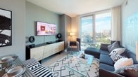 This single-floor residence is defined by its contemporary design and abundant natural light. Floor-to-ceiling windows in the living room and bedroom frame expansive views of the surrounding cityscape and tree canopy, creating a bright, airy atmosphere throughout. The living area offers a versatile layout with light wood-style flooring, neutral walls in soft sage tones, and ample space for both lounging and entertaining. A dedicated lounge nook positioned beside the large windows provides an ideal spot to enjoy the panoramic outlook.

The kitchen is outfitted with high-gloss white cabinetry, stainless steel appliances including a refrigerator, range, microwave, and dishwasher, complemented by a gray horizontal-striped tile backsplash and dark countertops. Track lighting overhead enhances functionality, while light hardwood flooring ties the space to the rest of the unit.

The bedroom features plush carpeting, generous closet access, and direct sightlines to the hallway and bathroom, reinforcing an open, connected feel. The full bathroom is finished with light wood-tone tile flooring, crisp white walls, a combined shower-tub with a curved curtain rod, and clean, modern fixtures throughout. The overall aesthetic is polished, cohesive, and well-suited to contemporary living.