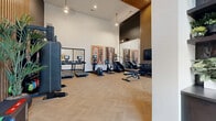 Located at 1425 Gallatin Pike N in Nashville, TN, this well-appointed fitness center offers a thoughtfully designed space for a comprehensive workout experience. The room is defined by soaring double-height ceilings and expansive floor-to-ceiling windows that flood the interior with natural light while framing views of the surrounding outdoor area, creating an open and energizing atmosphere.

The flooring features a striking light-toned herringbone pattern that runs throughout, complementing the contemporary aesthetic. Contrasting design elements — crisp white walls paired with bold dark accent walls adorned with vertical wood slat paneling — give the space a modern, upscale character. Curated artwork in gold-framed prints adds a polished, boutique-style touch.

The fitness center is fully equipped with a wide array of cardio machines, including treadmills, ellipticals, and rowing machines, each outfitted with individual digital displays. Strength training options include a multi-functional cable machine, a dumbbell rack, adjustable benches, medicine balls, and a BOSU trainer. Large wall-mounted mirrors enhance the sense of space and support proper form during workouts. A mounted flat-screen TV and integrated audio system complete the amenity. Lush potted tropical plants are strategically placed throughout, adding a refreshing natural element to the overall design.