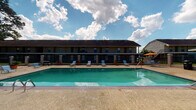 This outdoor pool area is a central amenity within a multi-building residential complex in Baton Rouge, Louisiana. The pool itself is a standout feature—large and rectangular with clear turquoise water, framed by a broad concrete deck lined with lounge chairs ideal for sunbathing. Bright orange umbrellas add pops of color and provide shaded seating near the pool's edge, while the entire pool zone is enclosed by a black iron fence with a gated entry, ensuring privacy and security.

Adjacent to the pool is a spacious covered pavilion supported by sturdy brick columns. This shaded structure offers a comfortable gathering space beneath its solid overhead canopy, with open sightlines toward the pool and surrounding green lawns. A decorative bench and waste receptacle furnish the pavilion, lending a practical, communal feel.

The support structure also houses a single-story utility or restroom building constructed of painted concrete block. Inside, gray doors with brass hardware lead to separate rooms, and the concrete floor and block walls give the space a utilitarian character. HVAC vents are integrated into the doors for climate control.

The broader complex features multi-story residential buildings with covered balconies, stairwells, and manicured green lawns connected by concrete walkways, all contributing to an organized and functional outdoor environment.