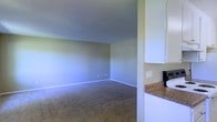 This single-story space offers a straightforward, functional layout highlighted by a spacious carpeted living area that flows openly toward the kitchen. The living area features neutral-toned walls with clean white baseboards, standard-height ceilings, and wall outlets conveniently positioned along the perimeter. The flooring throughout the main area consists of low-pile carpet in a warm beige tone, contributing to a comfortable and practical atmosphere. Adjacent to the living area, the kitchen is equipped with white upper cabinetry and granite-style countertops in earthy brown tones, providing ample storage and workspace. A light switch panel is visible near the kitchen entry, and the transition between spaces is open, enhancing the sense of connectivity within the unit. The overall color palette — soft greens on the walls paired with white trim and cabinetry — creates a cohesive, neutral aesthetic throughout. The space presents a clean, move-in ready environment well-suited for everyday living.