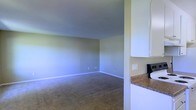 This single-story space offers a straightforward, functional layout highlighted by a spacious carpeted living area that flows openly toward the kitchen. The living area features neutral-toned walls with clean white baseboards, standard-height ceilings, and wall outlets conveniently positioned along the perimeter. The flooring throughout the main area consists of low-pile carpet in a warm beige tone, contributing to a comfortable and practical atmosphere. Adjacent to the living area, the kitchen is equipped with white upper cabinetry and granite-style countertops in earthy brown tones, providing ample storage and workspace. A light switch panel is visible near the kitchen entry, and the transition between spaces is open, enhancing the sense of connectivity within the unit. The overall color palette — soft greens on the walls paired with white trim and cabinetry — creates a cohesive, neutral aesthetic throughout. The space presents a clean, move-in ready environment well-suited for everyday living.