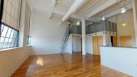 This industrial-style loft spans two levels, showcasing a striking open-concept design defined by soaring ceilings with exposed white-painted ductwork and structural columns anchoring the spacious main living area. Warm hardwood floors flow throughout, reflecting natural light that streams in through generously sized windows fitted with roller shades. A steel ladder leads up to the open loft level, bordered by a metal mesh railing, offering flexible additional living or sleeping space overlooking the main floor below.

The kitchen is thoughtfully appointed with light maple cabinetry, dark countertops, a slate-toned tile backsplash, and stainless steel appliances including a range, built-in microwave, refrigerator, and dishwasher. A checkered tile floor adds a classic touch to the culinary space. The bedroom features hardwood floors and large gridded windows that fill the room with natural light. A hallway with a warm wood-paneled door provides access between rooms. The full bathroom is finished with a light maple vanity, dark countertop, a tub-shower combination, and classic penny mosaic tile flooring, creating a clean, timeless aesthetic throughout.