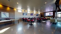 This single-floor sports bar and recreation lounge is designed to deliver an energetic yet comfortable social atmosphere. The main gathering area is expansive and open, featuring polished concrete floors that reflect ambient recessed lighting set into a clean, smooth ceiling. Light-toned tile-clad walls line the perimeter, punctuated by a row of narrow clerestory windows that allow natural light to filter in while maintaining a focused interior ambiance. Multiple flat-screen televisions are mounted throughout the space, ensuring unobstructed sightlines from every seat.

Seating throughout the main area consists of round high-top tables paired with red upholstered bar stools with black frames and gold footrests, creating a bold, sporty aesthetic. A large projection or display screen anchors one wall, complemented by a long bar-height counter ideal for group viewing. A separate lounge area offers cushioned armchairs for a more relaxed experience.

A dedicated game room adjacent to the main space features a full-size pool table and a ping-pong table, set against rich, dark wood-paneled walls warmly lit by accent lighting. Sports-themed artwork and photography are displayed throughout, reinforcing the venue's athletic identity. A transitional corridor with the same dark wood paneling leads to restrooms and utility areas, accented with a water fountain and decorative greenery.