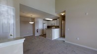 Located at 1515 Manley Road in West Chester, Pennsylvania, this single-floor residence offers a thoughtfully designed open-concept layout that seamlessly connects the main living and kitchen areas. The kitchen is appointed with white cabinetry, white appliances including a refrigerator, microwave, and electric range, and features a peninsula counter that provides both additional workspace and a natural divider between the kitchen and living space. A decorative pendant light hangs above, adding a touch of warmth to the area. The living space is finished with plush neutral carpeting that extends throughout, contributing to a comfortable and cohesive feel. Soaring ceilings elevate the sense of space, allowing the rooms to feel open and airy. The walls are finished in a soft, neutral beige tone that complements the overall palette. A hallway visible from the main area leads to additional rooms, suggesting a well-organized floor plan. The space is practical and move-in ready, with clean finishes and a functional design suited for everyday living in the heart of West Chester, PA.