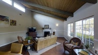 Located at 11355 Zapata Ave in San Diego, this single-story home makes a striking architectural statement with its soaring vaulted ceiling finished in warm wood planking and anchored by bold exposed beams. The living room serves as the heart of the home, showcasing the full grandeur of the ceiling height while maintaining an intimate, comfortable atmosphere. Rich hardwood flooring runs throughout, complementing the natural wood tones overhead and grounding the space with warmth and continuity. A classic fireplace with a white mantel serves as a focal point along one wall, adding both character and coziness to the room. Natural light filters in generously through multiple windows, including a high transom-style window that draws the eye upward and enhances the sense of openness. The living area is well-proportioned, offering ample space for relaxed seating arrangements centered around a woven rattan sofa and complementary wicker-style accent chairs, creating a casual yet refined aesthetic. Neutral wall tones provide a versatile backdrop throughout, allowing the architectural details and natural materials to take center stage. The overall ambiance is warm, airy, and relaxed — well-suited to the Southern California lifestyle.