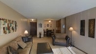 This single-floor residential space offers a welcoming and functional layout centered around an open-concept living and dining area. The living room is generously sized, featuring plush seating with leather-accented sofas, a cushioned ottoman, and warm ambient lighting provided by floor and table lamps. A textured ceiling adds character to the space, while neutral tan and brown tones throughout create a cohesive, understated aesthetic. Wall art and decorative accents lend a personal touch to the living area.

The dining space sits just beyond the living room, accommodating a dark wood dining table and chairs beneath a ceiling fan with integrated lighting — practical for year-round comfort. The carpeted floors extend throughout the main living and dining areas, contributing to the warm and relaxed atmosphere of the home.

Transitioning toward the back of the space, the kitchen is partially open to the dining area, featuring wood-toned cabinetry and a functional countertop layout suited for everyday cooking. The entry door, finished in dark wood, is visible from the living area, reinforcing the open sightlines that make this single-floor layout feel spacious and easy to navigate.