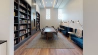 Located at 538-546 Broad St in Newark, NJ, this thoughtfully designed library space combines functionality with sophisticated style. The single-floor layout centers around a generous wood-topped conference table surrounded by upholstered gray chairs, making it well-suited for meetings, collaboration, or focused study sessions. Built-in, floor-to-ceiling dark charcoal bookshelves line the walls and are fully stocked with a diverse collection of books, lending the space a rich, scholarly atmosphere. A decorative gilt-framed mirror is integrated into the shelving, adding an elegant architectural accent. Along one wall, a row of tufted teal velvet sofas and lounge chairs provides a comfortable alternative seating area, accented by brass articulating wall sconces that cast warm, directional light. Tall, narrow arched windows allow natural light to filter in, highlighting the soaring ceiling height. The warm hardwood floors throughout are complemented by a neutral area rug beneath the conference table, adding texture and definition to the space. A small wet bar with a white subway tile backsplash, dark countertop, and sink is conveniently tucked near the entrance, and double glass doors provide easy access. The overall ambiance is polished, inviting, and purposefully curated.