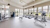 Located at 105 Exchange Circle in Asheville, NC, this fitness center offers a thoughtfully designed exercise facility spread across a single level. The space is divided into two distinct areas: a spacious main gym floor and a dedicated spin studio, together providing a comprehensive workout environment.

The main gym area is architecturally distinguished by soaring ceilings with a coffered, shiplap-clad design and pendant lighting that lends a warm, upscale ambiance. A generous bank of large windows lines the walls, flooding the floor with natural light while offering views of the surrounding greenery. The durable rubber-composite flooring provides a safe, functional surface throughout. Equipment includes multiple treadmills, elliptical machines, a cable machine, strength stations, a full dumbbell rack with benches, and various cable attachments—catering to both cardio and strength training needs. Wall-mounted flat-screen TVs keep users entertained during workouts.

The adjacent spin studio features hardwood-style flooring, a vibrant blue accent wall, floor-to-ceiling mirrors, and high windows that bring in additional natural light. A row of stationary spin bikes lines the room, complemented by wall-mounted televisions for guided or independent cycling sessions.

Together, these two zones create a well-rounded, inviting fitness facility suited for a wide range of exercise routines.