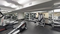 This fitness center is a comprehensively outfitted facility designed to support a full range of workout routines. The space is finished with durable dark rubber flooring throughout, providing a professional and practical surface suited for both cardio and strength training activities. Large floor-to-ceiling mirrors line the walls, visually expanding the room and allowing users to monitor their form during exercise. Overhead, recessed and flush-mounted lighting ensures the space is consistently and brightly illuminated.

The cardio zone features multiple treadmills and elliptical trainers from Life Fitness, each equipped with individual display screens for tracking workout metrics. A stair climber adds further variety to the cardio offerings. The strength training area is well-stocked with a full rack of dumbbells in graduated weights, a dedicated weight bench, and a cable/resistance machine, catering to users at all fitness levels.

A standout feature of the facility is a glass-enclosed steam room or sauna, accessible directly from the main floor and finished with clean white tile walls. Stainless steel waste receptacles and sanitation dispensers are thoughtfully placed throughout. The overall ambiance is modern and functional, reflecting a well-maintained, amenity-grade fitness environment.