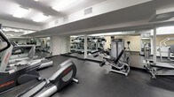 This fitness center is a comprehensively outfitted facility designed to support a full range of workout routines. The space is finished with durable dark rubber flooring throughout, providing a professional and practical surface suited for both cardio and strength training activities. Large floor-to-ceiling mirrors line the walls, visually expanding the room and allowing users to monitor their form during exercise. Overhead, recessed and flush-mounted lighting ensures the space is consistently and brightly illuminated.

The cardio zone features multiple treadmills and elliptical trainers from Life Fitness, each equipped with individual display screens for tracking workout metrics. A stair climber adds further variety to the cardio offerings. The strength training area is well-stocked with a full rack of dumbbells in graduated weights, a dedicated weight bench, and a cable/resistance machine, catering to users at all fitness levels.

A standout feature of the facility is a glass-enclosed steam room or sauna, accessible directly from the main floor and finished with clean white tile walls. Stainless steel waste receptacles and sanitation dispensers are thoughtfully placed throughout. The overall ambiance is modern and functional, reflecting a well-maintained, amenity-grade fitness environment.