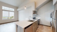 Located at 1217 Harris Ave, Unit 309 in Bellingham, WA, this modern two-story unit offers a well-designed layout spread across two levels. The main floor is anchored by an open-concept kitchen and living area, where light wood-tone flooring and neutral walls create a clean, contemporary aesthetic. The kitchen features a large island with a white quartz countertop, stainless steel appliances including an electric range with a wall-mounted hood, dishwasher, and a deep single-basin sink. A stylish pendant light hangs above the island, and a stacked washer and dryer unit is conveniently integrated into the kitchen area. Soaring ceilings amplify the sense of openness throughout the main level. The living areas are bathed in natural light from oversized windows fitted with transom panels, framing striking views of downtown Bellingham and Bellingham Bay beyond. The bedroom includes a sliding-door closet and maintains the same light flooring and high ceilings found throughout the unit. The full bathroom offers a single-sink vanity with quartz countertop, taupe cabinetry, a walk-in shower, and warm wood-look tile flooring. The overall ambiance is bright, clean, and modern, with thoughtful finishes throughout.
