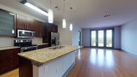 This well-appointed single-floor residence offers a thoughtfully designed open-concept layout that seamlessly connects the kitchen and living area. The kitchen is a standout feature, showcasing rich dark-wood cabinetry — including glass-front upper cabinets — paired with granite countertops, a subway tile backsplash, and a large central island with an integrated sink. Pendant lighting above the island adds a refined touch, while stainless steel appliances, including a side-by-side refrigerator, range, and microwave, complete the space. Warm hardwood flooring flows throughout the kitchen and expansive living area, which is filled with natural light from large double doors that open to an outdoor balcony, offering views of the surrounding urban landscape. The bedrooms feature plush carpeting, ceiling fans, and generous windows with blinds that bring in ample daylight. Two full bathrooms maintain a consistent design language with light tile flooring, tiled tub-shower combinations accented by dark tile borders, and dark wood cabinetry. A dedicated in-unit laundry area with a full-size washer and dryer adds everyday convenience. The overall aesthetic is modern and cohesive, with neutral tones and quality finishes throughout.