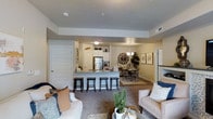 This single-floor residence offers an open-concept design that integrates the kitchen, dining, and living areas into one cohesive and functional space. The kitchen is outfitted with white cabinetry, granite countertops, and stainless steel appliances, including a full-size refrigerator. A breakfast bar with seating for three provides a casual dining option and serves as a natural divider between the kitchen and the main living areas. The dining area sits just beyond the kitchen and features a chandelier overhead, adding a refined touch to the space. The living room is carpeted and generously sized, furnished with comfortable seating and anchored by a glass-top coffee table. Tray ceilings add architectural depth and visual interest throughout the common areas. Interior doors are painted white and trimmed with clean molding, contributing to a bright and cohesive aesthetic. The overall ambiance is warm and inviting, with a neutral color palette that enhances the sense of openness. The layout flows naturally from one area to the next, making the space feel both practical and welcoming for everyday living.