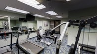 This fitness center offers a functional and practical workout environment housed on a single floor. The room is furnished with a selection of cardio machines, including treadmills and a recumbent bike, alongside strength and resistance training equipment. Mirrored walls line one side of the room, enhancing the sense of space while allowing users to monitor their form during exercise. The ceiling features recessed panel lighting that provides consistent, even illumination throughout the room. The floor is covered in a patterned carpet in deep blue tones, adding a subtle decorative element to the otherwise utilitarian space. A wall-mounted flat-screen television is positioned for easy viewing from the cardio equipment. A glass-paneled door provides access to and from the room, contributing to an open feel. Motivational signage is displayed on the walls, contributing to the overall fitness-focused atmosphere. The layout is efficient, positioning equipment to maximize usable floor space while maintaining comfortable circulation pathways between machines.