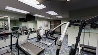This fitness center offers a functional and practical workout environment housed on a single floor. The room is furnished with a selection of cardio machines, including treadmills and a recumbent bike, alongside strength and resistance training equipment. Mirrored walls line one side of the room, enhancing the sense of space while allowing users to monitor their form during exercise. The ceiling features recessed panel lighting that provides consistent, even illumination throughout the room. The floor is covered in a patterned carpet in deep blue tones, adding a subtle decorative element to the otherwise utilitarian space. A wall-mounted flat-screen television is positioned for easy viewing from the cardio equipment. A glass-paneled door provides access to and from the room, contributing to an open feel. Motivational signage is displayed on the walls, contributing to the overall fitness-focused atmosphere. The layout is efficient, positioning equipment to maximize usable floor space while maintaining comfortable circulation pathways between machines.