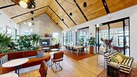 Located at 1 Samuel Peterson Dr in Titusville, NJ, this thoughtfully designed single-floor clubhouse offers a generous and versatile communal environment spanning a substantial open footprint. The architectural centerpiece is a dramatic vaulted ceiling clad in warm natural wood planks, supported by bold black steel beams that add an industrial contrast to the organic warmth of the space. Light oak hardwood flooring runs throughout, unifying the various zones seamlessly.

The main lounge area features a large tiled fireplace, lush built-in planters overflowing with greenery, and curved cognac leather banquette seating accented by black-and-white checkered cushions. A separate seating zone offers plush sectional sofas, layered with colorful throw pillows atop richly patterned area rugs. A dedicated dining and co-working area provides ample long tables with mixed seating for collaborative use.

A bar-height kitchen station is finished with striking floor-to-ceiling green mosaic tile, a sleek countertop, and bar stools — ideal for casual entertaining. A recreation zone includes a full-sized pool table beneath a sculptural gold chandelier. Floor-to-ceiling black-framed windows and multiple sets of glass doors flood every corner with natural light while offering direct views and access to the surrounding outdoor areas.