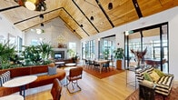 Located at 1 Samuel Peterson Dr in Titusville, NJ, this thoughtfully designed single-floor clubhouse offers a generous and versatile communal environment spanning a substantial open footprint. The architectural centerpiece is a dramatic vaulted ceiling clad in warm natural wood planks, supported by bold black steel beams that add an industrial contrast to the organic warmth of the space. Light oak hardwood flooring runs throughout, unifying the various zones seamlessly.

The main lounge area features a large tiled fireplace, lush built-in planters overflowing with greenery, and curved cognac leather banquette seating accented by black-and-white checkered cushions. A separate seating zone offers plush sectional sofas, layered with colorful throw pillows atop richly patterned area rugs. A dedicated dining and co-working area provides ample long tables with mixed seating for collaborative use.

A bar-height kitchen station is finished with striking floor-to-ceiling green mosaic tile, a sleek countertop, and bar stools — ideal for casual entertaining. A recreation zone includes a full-sized pool table beneath a sculptural gold chandelier. Floor-to-ceiling black-framed windows and multiple sets of glass doors flood every corner with natural light while offering direct views and access to the surrounding outdoor areas.