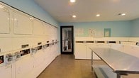 This shared laundry facility is a single-floor space designed with practicality and efficiency in mind. The room is finished with light blue walls and recessed ceiling lighting, creating a bright and clean environment for residents. Along the left wall, a row of white front-load dryers is stacked in pairs, offering ample drying capacity. Opposite them, a lineup of top-load washing machines provides convenient access for laundry loads of various sizes. A stainless steel folding table is positioned along one wall, giving users a dedicated surface for sorting and folding garments. The tile flooring is durable and easy to maintain, well-suited to the demands of a high-use utility space. A glass-paneled door provides access to an adjacent area, adding a sense of openness to the room. Informational notices and usage guidelines are posted on the walls, ensuring users have easy access to important instructions. Overall, the space is thoughtfully organized to maximize functionality, making it a practical and accessible amenity for its users.