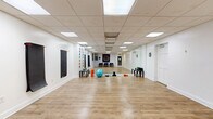 Located at 104 W Franklin St in Richmond, Virginia, this single-floor wellness studio offers a thoughtfully designed open layout ideal for yoga, fitness, or holistic practices. The space is defined by warm light wood laminate flooring that extends throughout, creating a clean and inviting foundation for movement-based activities.

The main studio area is bright and open, illuminated by a grid of recessed panel ceiling lights that provide consistent, even lighting throughout. Crisp white walls keep the environment feeling fresh and expansive. Two wall-mounted black exercise/yoga mats hang neatly on one wall, offering convenient storage and easy access during sessions. A full-length mirror along one wall enhances the sense of depth and supports movement practice and form correction.

Decorative elements reflect a mindful, wellness-focused aesthetic — a colorful chakra symbol display lines one wall, while a large intricate mandala tapestry serves as a striking focal point in the seating area. A map of Richmond and additional spiritual wall art add personality to the space.

A small seating area with blue upholstered chairs provides a welcoming reception or rest zone. Fitness props including a stability ball, resistance bands, and exercise mats are present throughout the studio floor. The space also features a separate entry door and utility access, making it a fully functional, self-contained studio environment.