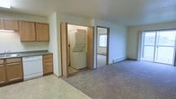 This single-floor residential unit offers a practical and functional layout designed for comfortable everyday living. The kitchen features light oak cabinetry, a dark countertop, and tile flooring, providing a clean and efficient workspace. Adjacent to the kitchen is a dedicated laundry closet housing a stacked washer and dryer unit, adding significant in-unit convenience. The open living area is finished with wall-to-wall carpet and is served by baseboard heating, ensuring warmth throughout the space. A doorway leads to an additional carpeted room, also equipped with baseboard heating and a window that allows natural light to filter in. The walls throughout are neutral in tone, contributing to a bright and understated interior. The layout connects the functional zones of the home in a straightforward and accessible manner, making efficient use of the available floor space. Overall, the unit presents a no-frills, move-in-ready environment well suited for those seeking practical amenities and a simple, cohesive living space on a single level.