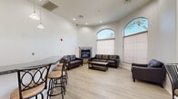 This well-appointed single-floor communal space offers a generous, open layout designed for comfort and social gathering. The room is anchored by soaring ceilings that create an airy, expansive atmosphere, complemented by crisp white walls and light hardwood-style flooring throughout.

The lounge area is a standout feature, centered around a stone-surround fireplace that provides a warm, inviting focal point. Comfortable leather seating is arranged to encourage conversation, while large arched windows flood the space with abundant natural light and frame views of the surrounding outdoor landscape. Cellular shades offer adjustable privacy without sacrificing brightness.

A dedicated dining nook near the entrance features a high-top table with bar-height seating, perfect for casual gatherings. Sleek black French doors open directly to an outdoor courtyard area, seamlessly connecting the interior to the exterior environment.

The kitchenette is a practical and stylish addition, boasting warm wood cabinetry, dark granite countertops, and black appliances including a refrigerator and microwave. A breakfast bar with bar stools provides additional seating and serves as a natural divider between the kitchen and lounge areas. Pendant lighting overhead adds a refined finishing touch. Arched architectural details and recessed lighting further elevate the overall design of this versatile community space.