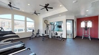 This fitness center is designed to provide a comprehensive workout experience within a single-level space. The room is divided into two connected zones, each with its own distinct character while maintaining a cohesive overall layout.

The main workout area features durable rubber flooring throughout, offering both safety and comfort during exercise. Multiple ceiling fans are installed across both zones, ensuring consistent air circulation during workouts. Recessed lighting supplements the abundant natural light that streams in through large arched and rectangular windows, providing clear views to the exterior and keeping the space bright and inviting.

One zone showcases a tray ceiling detail, lending architectural interest to the space. A large wall mirror expands the visual depth of the room and supports exercise form-checking. This area houses a multi-station cable and weight machine, a stationary bike, and additional strength equipment, along with free weights accessible along the perimeter.

The second zone is anchored by a bold red accent wall, creating an energizing atmosphere. This area is dedicated to cardio equipment, including multiple treadmills and elliptical trainers positioned facing the windows. A wall clock keeps exercisers on schedule. Together, both zones create a well-rounded, functional fitness facility.