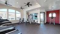 This fitness center is designed to provide a comprehensive workout experience within a single-level space. The room is divided into two connected zones, each with its own distinct character while maintaining a cohesive overall layout.

The main workout area features durable rubber flooring throughout, offering both safety and comfort during exercise. Multiple ceiling fans are installed across both zones, ensuring consistent air circulation during workouts. Recessed lighting supplements the abundant natural light that streams in through large arched and rectangular windows, providing clear views to the exterior and keeping the space bright and inviting.

One zone showcases a tray ceiling detail, lending architectural interest to the space. A large wall mirror expands the visual depth of the room and supports exercise form-checking. This area houses a multi-station cable and weight machine, a stationary bike, and additional strength equipment, along with free weights accessible along the perimeter.

The second zone is anchored by a bold red accent wall, creating an energizing atmosphere. This area is dedicated to cardio equipment, including multiple treadmills and elliptical trainers positioned facing the windows. A wall clock keeps exercisers on schedule. Together, both zones create a well-rounded, functional fitness facility.