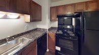 This single-floor residence offers a well-organized layout with distinct living, dining, kitchen, bedroom, and bathroom areas. The living and dining spaces flow together, with the dining area featuring wood-look flooring and a pendant light fixture overhead, while the adjacent living area is finished with plush carpet. A partial wall separates these areas from the kitchen, creating a semi-open feel.

The kitchen is equipped with rich, warm-toned wood cabinetry extending to the ceiling, dark granite-style countertops, a stainless steel sink, and black appliances including an electric range, over-the-range microwave, and dishwasher. A hanging pendant light provides focused illumination over the workspace.

The bedrooms are carpeted and feature natural light through large windows dressed with vertical blinds. Each bedroom includes a built-in closet with bifold doors offering organized storage space. Textured ceilings are consistent throughout the unit.

The bathrooms feature dark marble-style countertops with warm wood vanity cabinetry, single-basin sinks, and mirrored medicine cabinets above. Tile flooring distinguishes the bathroom spaces from the carpeted rooms nearby. Overall, the space presents a cohesive, functional design with consistent finishes throughout.