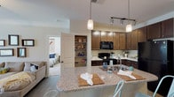 Located in Clayton, North Carolina, this thoughtfully designed single-floor residence offers a comfortable and functional layout ideal for everyday living. The heart of the home is an open-concept kitchen equipped with rich dark cabinetry, granite countertops, black appliances including a range and built-in microwave, and a spacious island with pendant lighting overhead. A dedicated walk-in pantry provides generous dry storage. The kitchen flows seamlessly into a dining area finished with wood-look flooring and crown molding, lending a polished, classic feel to mealtimes. A cozy living area with neutral tones and hardwood-style floors offers a relaxed gathering space. The bedrooms are carpeted and well-lit by large windows that invite abundant natural light, each featuring ceiling fans for year-round comfort. A spacious walk-in closet with wire shelving serves the primary bedroom. The bathroom is outfitted with a granite-topped dark vanity, wood-look tile flooring, and a full tub-shower combination. A dedicated interior laundry room accommodates a full-size washer and dryer with overhead shelving for added utility. Throughout, the home maintains a warm, neutral palette complemented by crown molding and clean architectural lines.