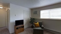 This single-floor space offers a straightforward and functional layout designed for comfortable everyday living. The living area is carpeted and features a neutral color palette with white walls and a textured ceiling that adds a subtle visual element to the room. A wall-mounted air conditioning unit provides climate control, while baseboard heating along the wall ensures warmth during cooler months. A window fitted with horizontal blinds allows natural light to brighten the space, creating a welcoming atmosphere. The room comfortably accommodates a plush armchair with a decorative yellow accent pillow, providing a relaxed seating area, alongside a wooden media console supporting a flat-screen television. A potted indoor plant adds a touch of greenery, softening the utilitarian elements of the room. A six-panel interior door leads to an adjacent area, contributing to the efficient use of the floor plan. Overall, the space presents a clean, no-frills environment that balances practicality with comfort, making it well-suited for day-to-day residential use.
