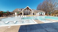 This single-story community amenity center is designed to deliver a resort-like experience for its residents. At the heart of the outdoor space is a large, sparkling swimming pool with a zero-entry wading section, multiple in-pool seating areas, and accessibility features including a pool lift. The stamped concrete pool deck provides generous space for lounging, lined with chairs and shaded by green market umbrellas. The clubhouse building itself features a striking gabled roofline with board-and-batten siding, large multi-pane windows, and French doors that open directly to the pool area, allowing seamless indoor-outdoor flow. A covered outdoor living area extends from the building and showcases a warm wood-paneled ceiling with ceiling fans, a wall-mounted television, a built-in fireplace framed in white shiplap with a brick surround, and comfortable seating arrangements. An outdoor kitchen equipped with a built-in grill and brick surround sits adjacent to the covered patio, ideal for gatherings and events. The space is enclosed by a black iron fence and surrounded by mature trees and manicured landscaping, creating a private and inviting atmosphere that balances elegance with everyday functionality.