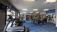 Located at 1000 San Marcos St in Austin's vibrant 78702 zip code, this single-floor fitness center is a comprehensive workout facility designed to support a wide range of exercise routines. The space features a bold, energetic aesthetic with blue patterned carpet throughout, gray walls accented with motivational signage — including "Results" and "Challenge to Change" — and bright recessed and panel lighting that keeps the entire floor well-illuminated.

The main workout area is generously proportioned, housing a full suite of True-brand cardio equipment including treadmills, ellipticals, and a StairMaster, as well as a rowing machine. A dedicated strength training zone features multiple weight racks, barbells, dumbbells, and cable machines, offering ample options for resistance training. Floor-to-ceiling mirrors line portions of the walls, enhancing the sense of space and allowing users to monitor their form.

A separate cycling studio accommodates multiple stationary bikes and benefits from large glass doors that open toward an outdoor pool area, filling the room with natural light and views of the surrounding greenery. The main cardio area similarly features expansive windows overlooking the pool, creating a bright and inviting atmosphere. Mounted flat-screen TVs are positioned throughout the space for entertainment during workouts, and a colorful wall-mounted accessory station rounds out the functional offerings.