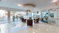 Located at 8255 Walnut Hill Lane in Dallas, Texas, this single-floor amenity space offers a thoughtfully designed environment with a variety of distinct areas, each finished with high-end materials and a cohesive contemporary aesthetic.

The main lounge area is anchored by large floor-to-ceiling glass walls that flood the space with natural light and provide direct views of an outdoor courtyard and pool area. Polished large-format white tile flooring runs consistently throughout, reinforcing the clean, modern feel. A central seating vignette with leather armchairs surrounds a sculptural, curved sofa enclosure draped in elegant chain curtains, creating an intimate focal point within the open layout.

A dedicated media lounge offers a dramatically different atmosphere, featuring dark walls adorned with an intricate geometric patterned wallcovering, a matte black ceiling, and a generously sized leather sectional sofa, designed for an immersive entertainment experience.

The wet bar area is outfitted with rich mosaic tile cladding, dark countertops, built-in cabinetry, a wall-mounted television, and a sink — seamlessly connecting to a bar-height dining area, ideal for social gatherings.

Glass-partitioned rooms along a bright corridor provide additional flexible spaces, while curated artwork along the walls contributes to the gallery-like character of the overall environment.