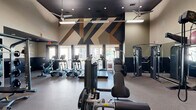 Located at 18235 Bulverde Rd in San Antonio, Texas, this fitness center offers a generously proportioned, single-floor exercise space designed to support a full range of workout routines. The room features soaring ceilings accented by a dark tray ceiling detail, ceiling fans for air circulation, and bright overhead lighting that ensures excellent visibility throughout.

A striking geometric mural in dark navy, tan, and black tones dominates one wall, adding a bold, modern aesthetic to the space. Large windows along the same wall flood the area with natural light and provide views of the outdoor surroundings, creating an energizing atmosphere for workouts.

The fitness center is comprehensively equipped with a variety of TRUE-brand cardio machines, including treadmills, ellipticals, and stair climbers, all positioned to take advantage of the natural light and exterior views. The strength training section features a multi-station cable machine, a leg press/curl machine, a Smith machine, and a dedicated free weights area stocked with dumbbells, kettlebells, medicine balls, and stability balls organized on storage racks.

Full-length mirrors line one wall, visually expanding the space and allowing users to monitor their form. Durable, low-pile carpet flooring covers the exercise area, while a wall-mounted television adds convenience. The overall design balances functionality with a clean, modern aesthetic suited to an active community.