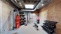 Located at 1400 S 2nd St in Wilmington, NC, this single-floor fitness space combines raw industrial character with a well-equipped workout environment. The room features striking exposed brick walls with a weathered, textured finish that lends an authentic, urban aesthetic. Overhead, exposed metal ductwork runs alongside track lighting and a central skylight, which draws in natural light and adds to the open, airy feel of the space despite its intimate footprint.

The gym is thoughtfully outfitted with a diverse range of equipment to support varied training styles. A heavy punching bag and battle ropes cater to combat and high-intensity interval training, while a multi-use storage rack holds medicine balls, resistance tools, and accessories. A flat utility bench sits centrally for strength and functional exercises, and a full dumbbell rack along the far wall provides options for progressive resistance training. Kettlebells and additional free weights round out the equipment selection.

The flooring is light-toned and low-pile, offering a clean contrast to the rugged brick surroundings. The overall ambiance is energetic and industrial, making this space well-suited for personal training, boutique fitness, or a private gym setting. Its architectural character and practical layout make it both visually compelling and highly functional.