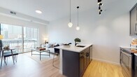 This contemporary single-floor residence showcases a thoughtfully designed open-concept layout that seamlessly connects the kitchen, dining, and living areas. The kitchen is a standout feature, equipped with dark cabinetry, stainless steel appliances, a hexagonal tile backsplash, and a large center island topped with a light quartz countertop. Stylish pendant lighting with geometric frosted globes and a matte black track system illuminates the space, while light hardwood flooring runs throughout, adding warmth and continuity. The living area is bright and airy, benefiting from a large sliding glass door that opens onto a private balcony offering sweeping city skyline views. This outdoor access floods the room with natural light and creates an inviting indoor-outdoor connection. The two bedrooms are calm and well-appointed, each featuring large windows that frame impressive urban vistas. The bathroom presents a clean, classic aesthetic with white penny tile flooring and matte black fixtures and hardware, complementing the modern design language found throughout the residence. The overall ambiance is one of refined urban living — contemporary, light-filled, and effortlessly connected to the city beyond.