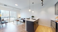 This contemporary single-floor residence showcases a thoughtfully designed open-concept layout that seamlessly connects the kitchen, dining, and living areas. The kitchen is a standout feature, equipped with dark cabinetry, stainless steel appliances, a hexagonal tile backsplash, and a large center island topped with a light quartz countertop. Stylish pendant lighting with geometric frosted globes and a matte black track system illuminates the space, while light hardwood flooring runs throughout, adding warmth and continuity. The living area is bright and airy, benefiting from a large sliding glass door that opens onto a private balcony offering sweeping city skyline views. This outdoor access floods the room with natural light and creates an inviting indoor-outdoor connection. The two bedrooms are calm and well-appointed, each featuring large windows that frame impressive urban vistas. The bathroom presents a clean, classic aesthetic with white penny tile flooring and matte black fixtures and hardware, complementing the modern design language found throughout the residence. The overall ambiance is one of refined urban living — contemporary, light-filled, and effortlessly connected to the city beyond.